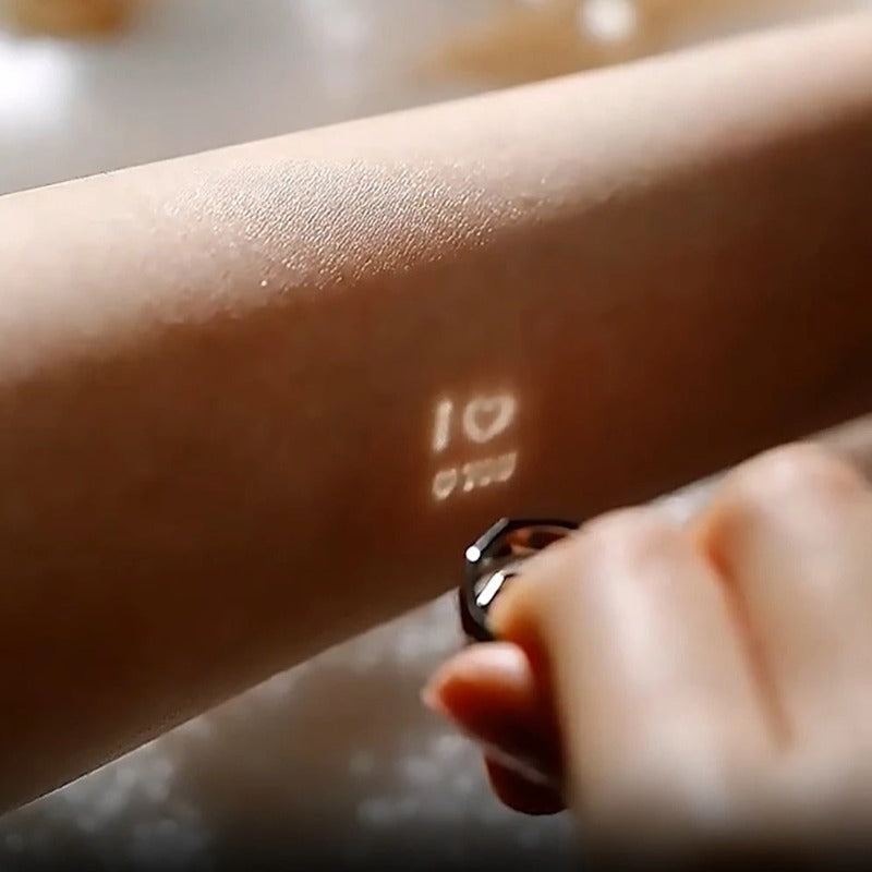 A hand holds an adjustable ring close to a forearm. The Jingqian S925 Sterling Silver Light Reflection Ring projects a small light that displays "I â¤ï¸ U" and another small symbol or text below it on the skin.