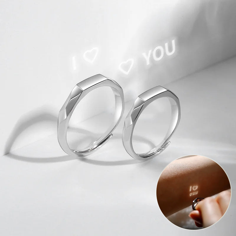 Two sterling silver rings with a flat, angular design are shown. One ring projects "I â™¥ YOU" in light onto a surface. An inset image displays a close-up of someone holding an adjustable Jingqian S925 Sterling Silver Light Reflection Ring and the same "I â™¥ YOU" message projected on their finger.
